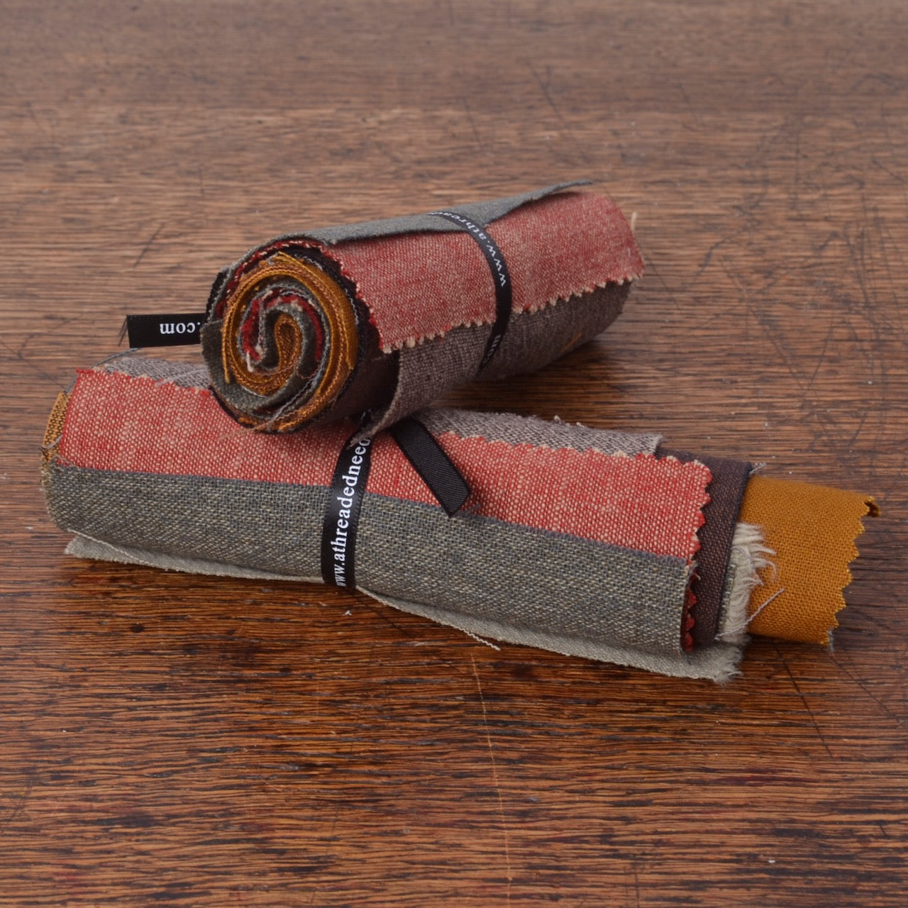 Two rolled fabric swatches with visible woven texture . The fabrics show muted red, grey, brown, mustard yellow, and green tones.