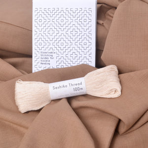 A skein of cream-colored Sashiko thread labeled 'Sashiko Thread 100m' resting on draped tan fabric, with a white instruction booklet featuring geometric stitching guides behind it.