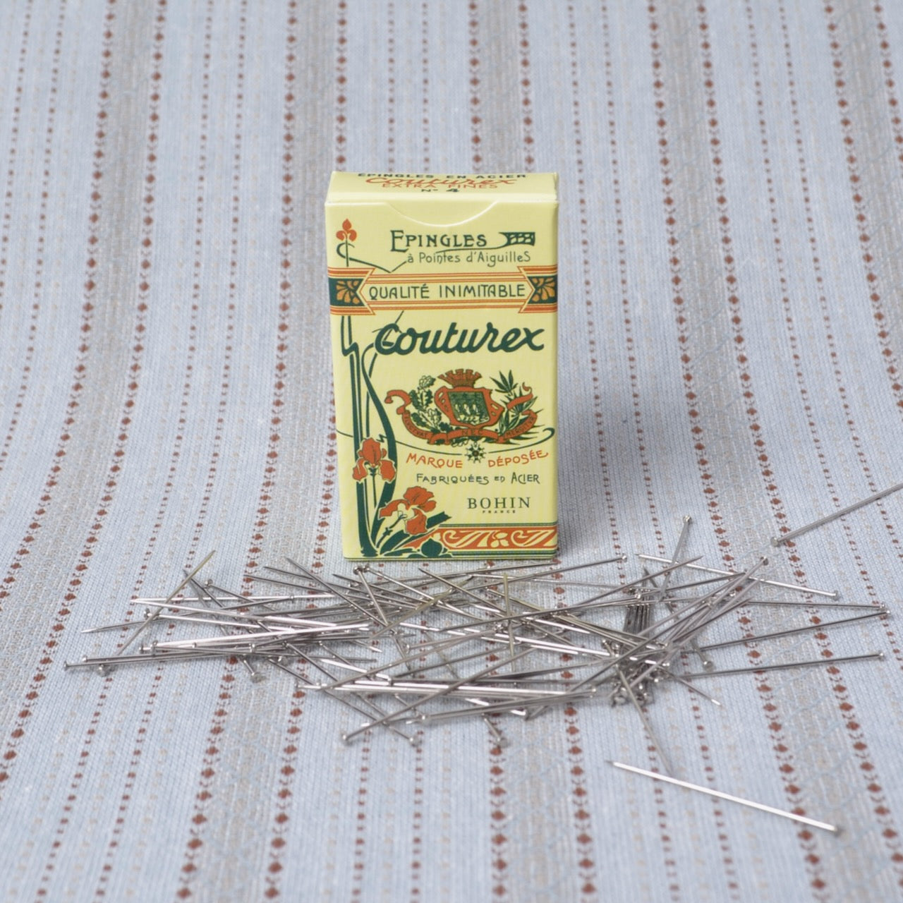 A vintage-style box of Couturex sewing pins by Eppingers stands on a light blue and white striped fabric surface, with several silver metal pins scattered in front of the box.