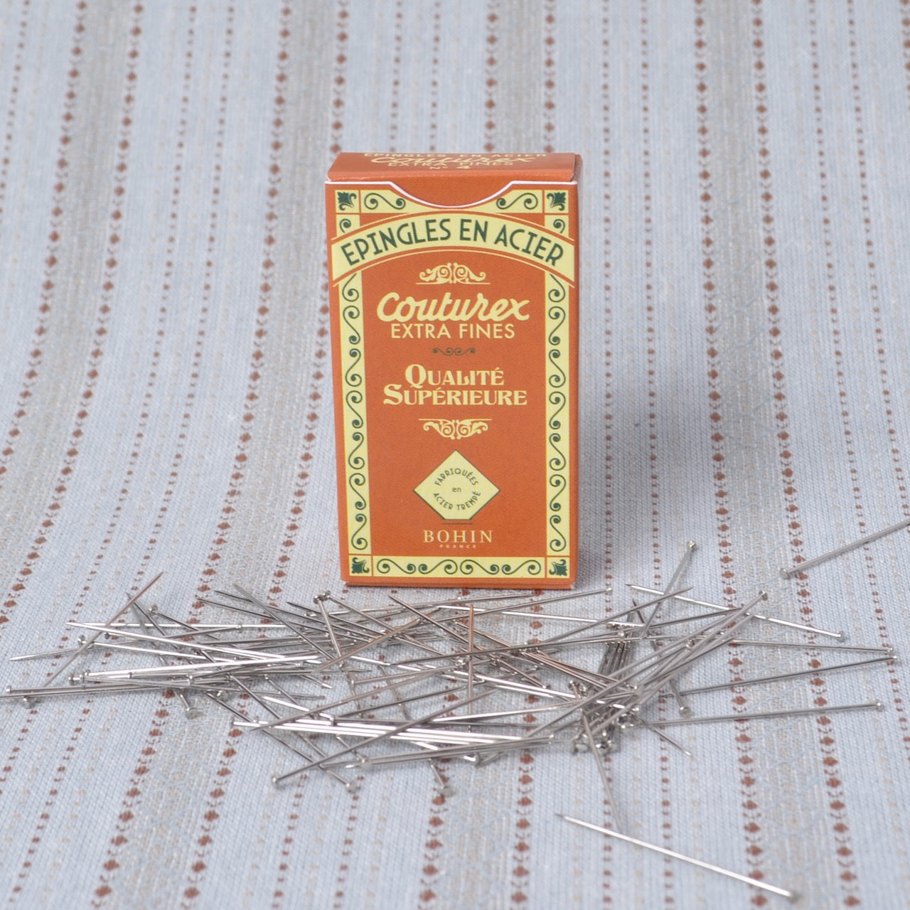 A box of Couturex extra fine sewing pins with several silver pins scattered in front on a light blue patterned fabric background.