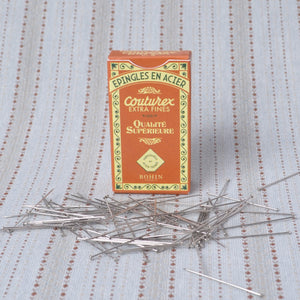 A box of Couturex extra fine sewing pins with several silver pins scattered in front on a light blue patterned fabric background.