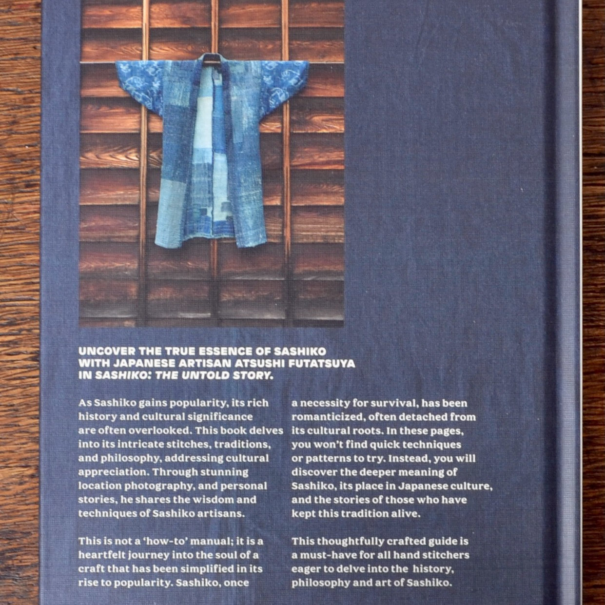 The back cover of a book titled 'Sashiko: The Untold Story' featuring a photograph of a blue-and-white patterned sashiko garment hanging on a wooden rack, set against a dark blue background with white text.