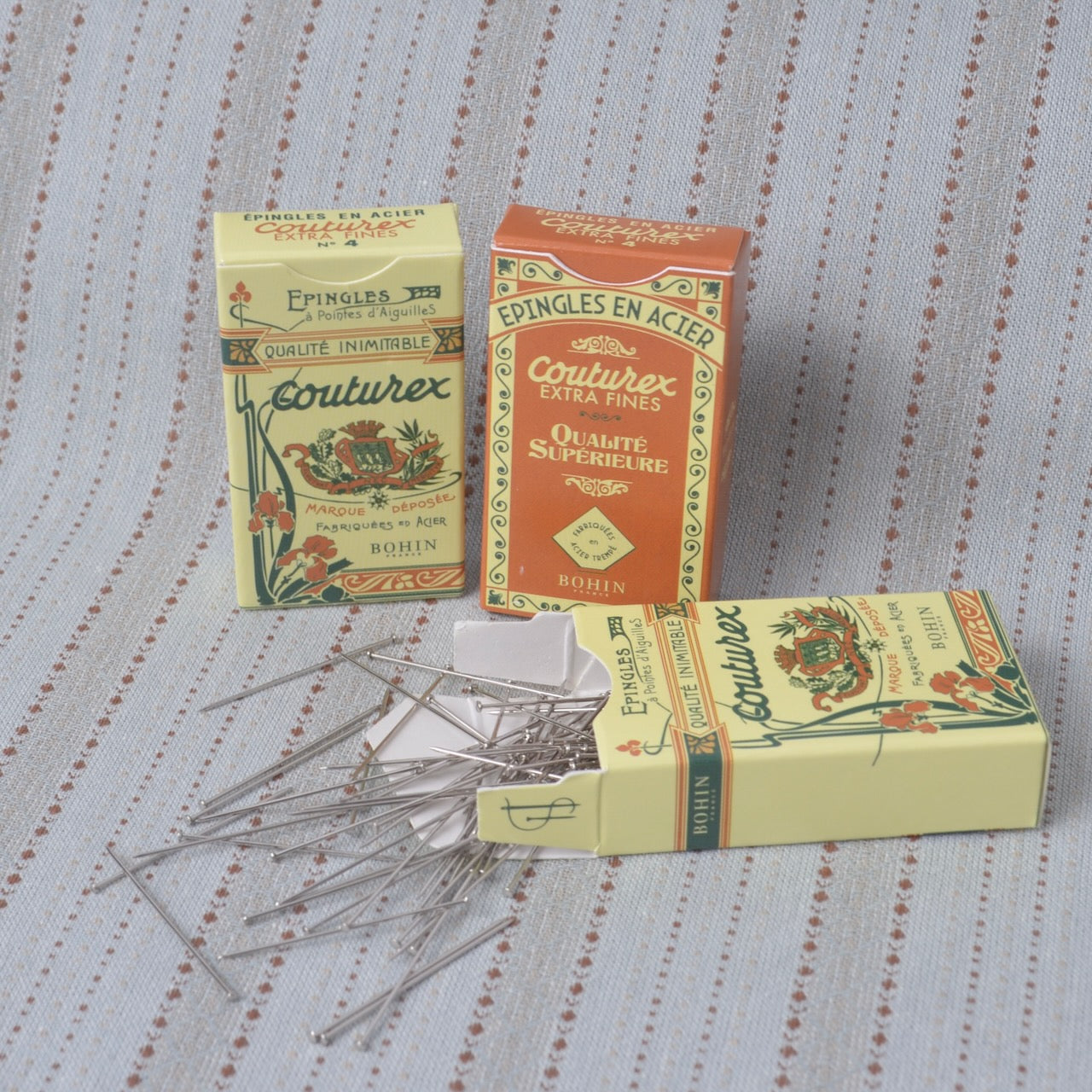 Three vintage boxes of Bouturex sewing pins arranged on a striped fabric background, with several silver pins spilled from the open box in the foreground.