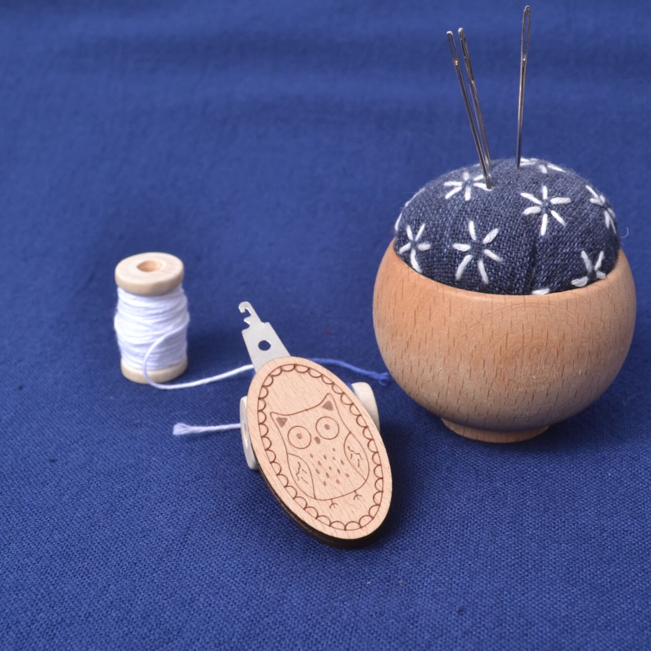 Wooden pincushion with floral pattern, spool of thread, and owl-themed bobbin on a blue fabric background